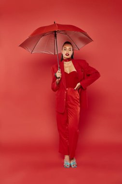 pretty african american woman in red pants and blazer standing under umbrella, hand on hip clipart