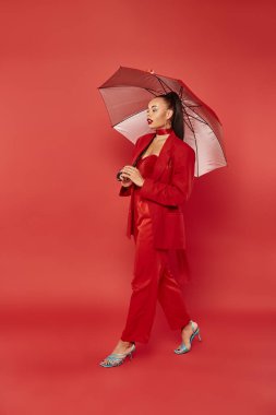 pretty african american woman in blazer and pants walking under umbrella on red backdrop clipart