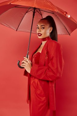 Kırmızı arka planda şemsiye altında duran ceketli ve pantolonlu güzel Afrikalı Amerikalı kadın.