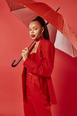 elegant african american woman in suit pants and blazer standing under umbrella on red backdrop clipart