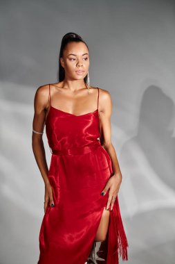 Kırmızı elbiseli çekici Afro-Amerikalı kadın aynalı gri arka planda poz veriyor.