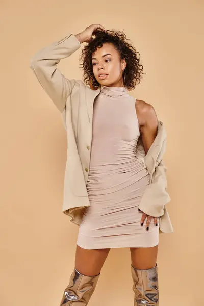 stock image beautiful african american woman adjusting curly hair and posing in stylish autumn attire on beige