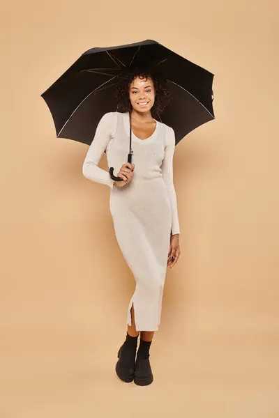 stock image happy african american woman in midi dress and boots standing under umbrella on beige, autumn look