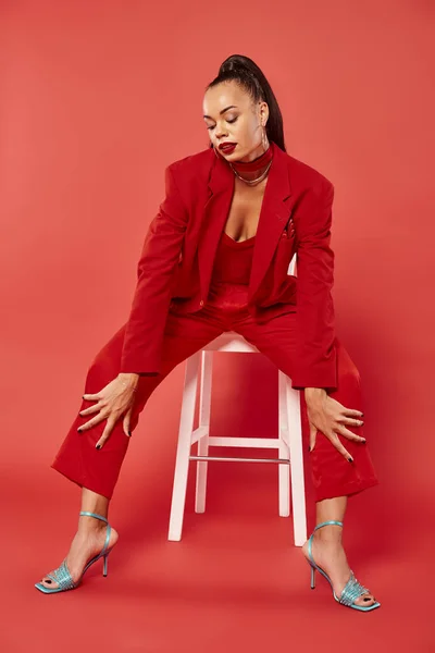 full length of elegant african american woman in suit and high heels sitting on white chair on red