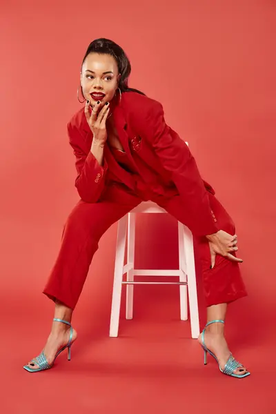 full length of elegant african american model in suit and high heels sitting on white chair on red