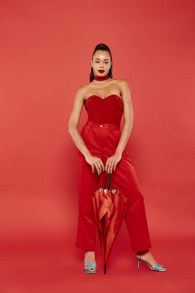 full length of charming african american model in red strapless top and pants posing with umbrella