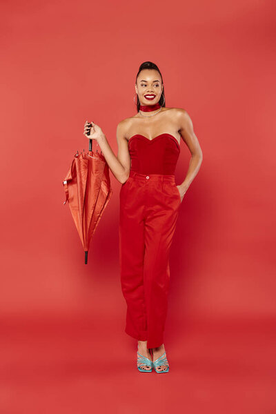 full length of happy african american model in red strapless top and pants posing with umbrella