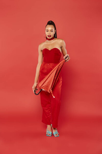 full length of brunette african american model in red strapless top and pants posing with umbrella