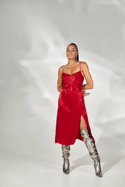 stock image full length, confident african american woman in red dress and silver boots posing on grey backdrop