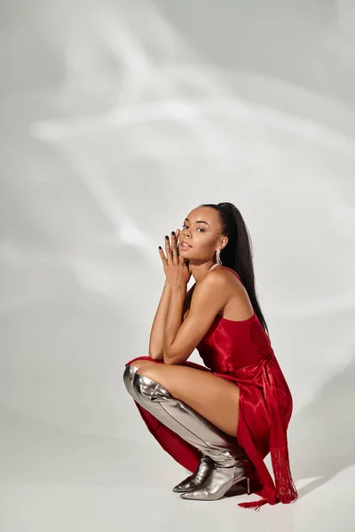 pretty african american woman in red midi dress and silver boots sitting on mirrored grey backdrop
