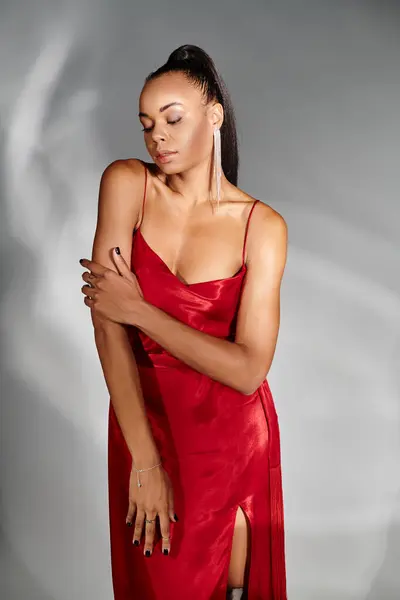 stock image glamorous african american woman in red dress looking down and posing on mirrored grey backdrop