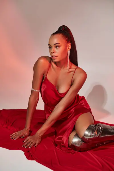 stock image young african american woman in red slip dress and silver boots sitting on fabric with grey backdrop