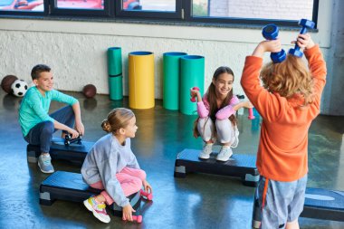 Dört neşeli çocuk spor ayakkabıları üzerinde dambıllarla poz veriyor ve neşeyle gülümsüyor, çocuk sporu.