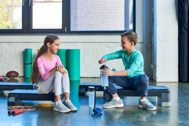 Spor giyimli neşeli ergen çocuklar su şişeleri ve spor ayakkabılarıyla spor basamaklarında oturuyorlar.