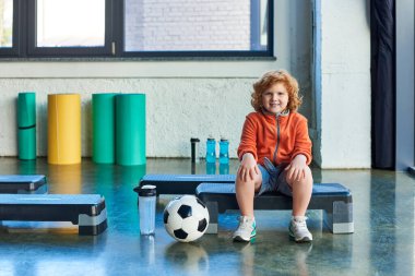 Futbol topu ve su şişesinin yanında oturan neşeli kızıl saçlı çocuk, çocuk sporu.
