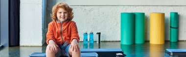 happy red haired boy sitting on fitness stepper smiling cheerfully at camera, child sport, banner