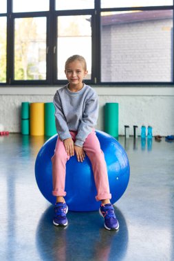cute preadolescent girl in sportswear sitting on fitness ball and looking at camera, child sport