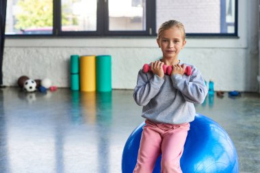 pretty blonde girl sitting on fitness ball and exercising with dumbbells, looking at camera, sport