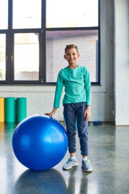 Mavi fitness topunun yanında durup kameraya bakan neşeli küçük çocuğun dikey görüntüsü.