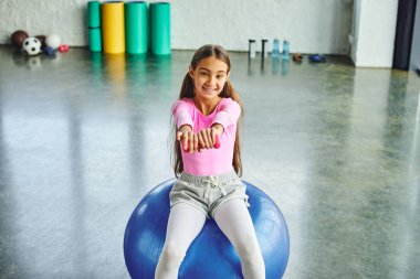 preadolescent cute girl in pink sportswear exercising with dumbbell on fitness ball, child sport clipart