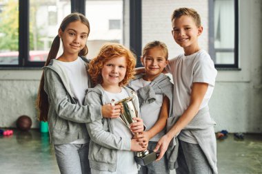 four joyous preadolescent children in grey sportswear posing with trophy in gym, child sport clipart