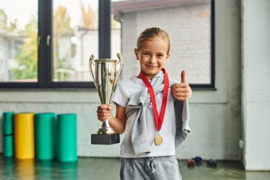 cheerful preadolescent girl with golden medal showing thumb up and holding trophy, child sport clipart