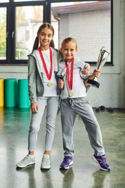 vertical shot of cute preadolescent girls with medals and trophy hugging and smiling at camera clipart