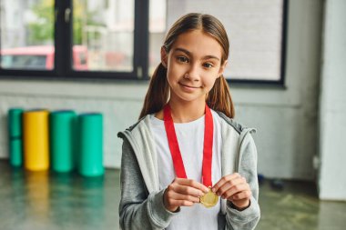smiley pretty girl with long hair holding golden medal and looking joyfully at camera, child sport clipart