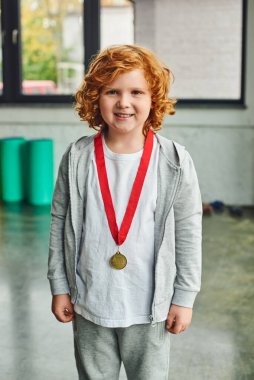 joyful red haired boy in sportswear with golden medal smiling sincerely at camera, child sport clipart