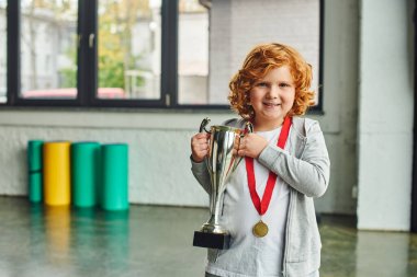 pretty red haired boy with golden medal holding huge trophy and smiling cheerfully at camera clipart