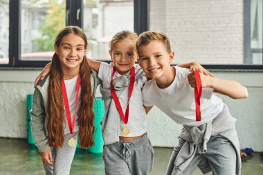 Altın madalyalı üç küçük arkadaş kameraya sarılıyor ve gülümsüyor, çocuk sporu