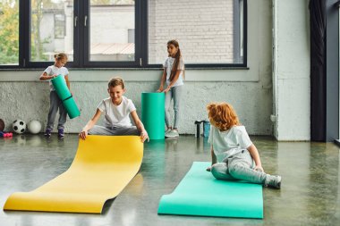 cheerful preadolescent children in sportswear unfolding fitness mats in gym, workout, child sport clipart