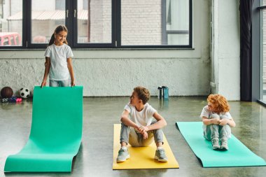 two preadolescent joyful boys smiling and looking at little cute girl unfolding fitness mat clipart