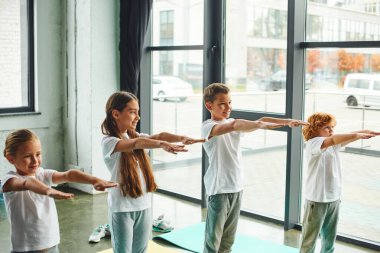Spor kıyafetleri içinde neşeli küçük çocuklar spor salonunda kollarını açıyorlar pencere önü ile, çocuk sporu