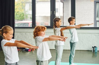 Beden eğitimi dersinde kollarını açıp spor minderlerine basan neşeli çocuklar.