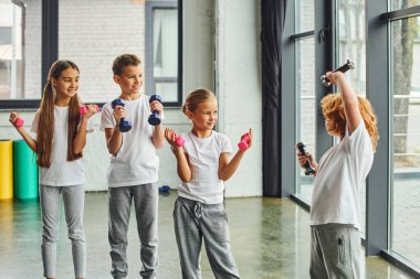 Neşeli ergenlik öncesi erkek ve kızlar spor salonunda halterleriyle egzersiz yapıyor, neşeyle gülümsüyor, spor yapıyor.