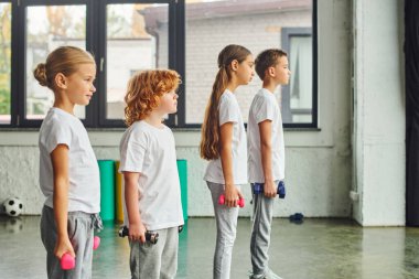 Erkek ve kız çocukları ellerinde dambıllarla spor salonunda poz veriyorlar, çocuk sporu.