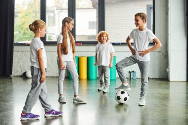 İki kız ve kızıl saçlı çocuk diğer çocuğa gülümsüyor bacağını futbol topuna koyuyor, çocuk sporu.