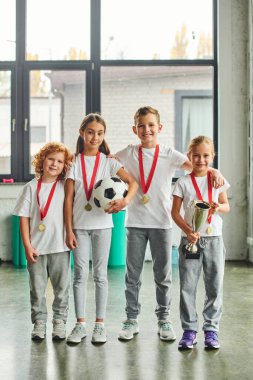 vertical shot of preadolescent cute children with golden medals posing with soccer ball and trophy clipart