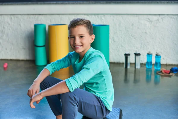 stock image cheerful adolescent boy sitting on fitness stepper and smiling sincerely at camera, child sport