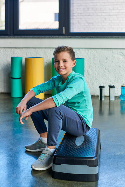 joyful boy in sportswear sitting on fitness stepper and smiling happily at camera, child sport