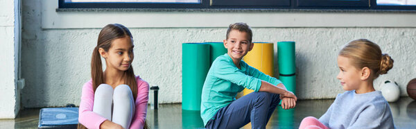 two pretty girls and boy in sportswear sitting in gym and smiling at each other, child sport