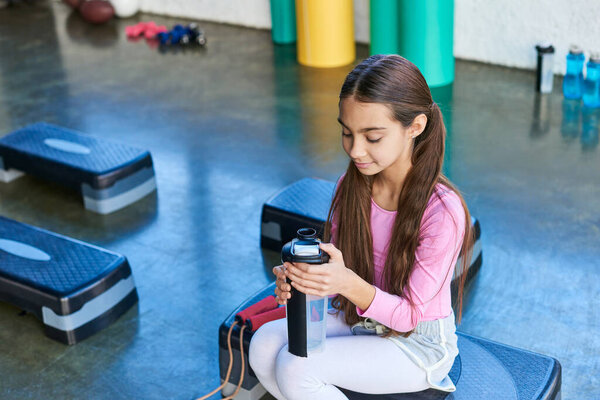 cheerful little girl holding water bottle sitting on fitness stepper with jump rope, child sport