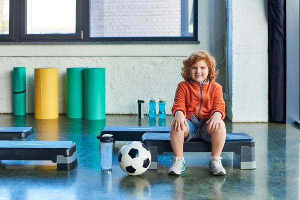 joyful red haired boy sitting on fitness stepper next to soccer ball and water bottle, child sport