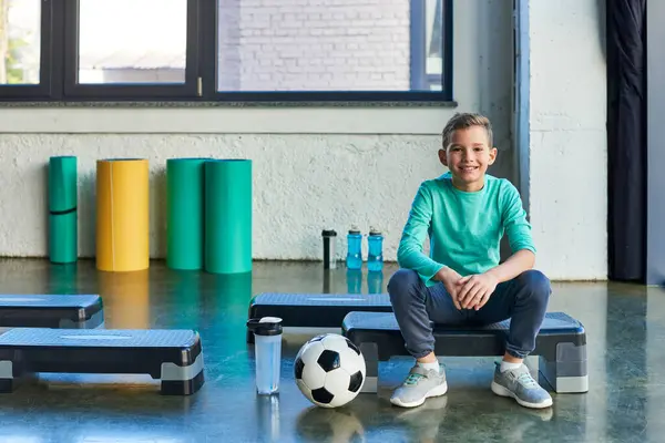 cheerful preadolescent boy on fitness stepper next to soccer ball and water bottles, child sport