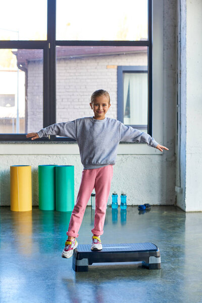 vertical shot of little cute girl stretching on fitness stepper and smiling at camera, child sport