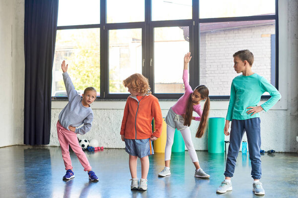 two little cute girls stretching in gym while boys looking attentively at them, child sport