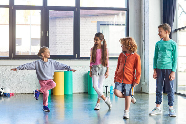 little blonde girl standing on one leg stretching in front of other cheerful kids, child sport