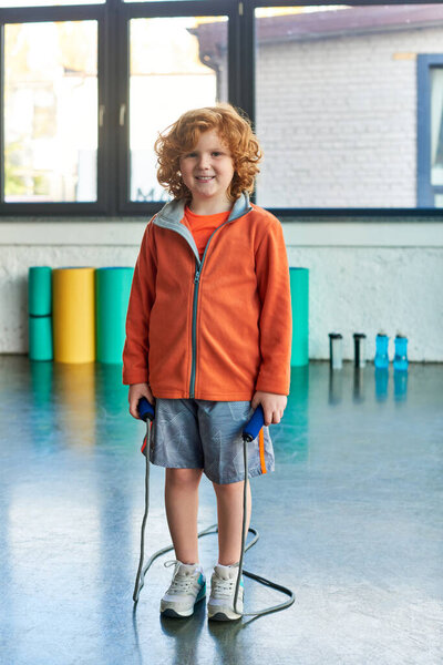 cheerful red haired boy posing with skipping rope and smiling joyfully at camera, child sport