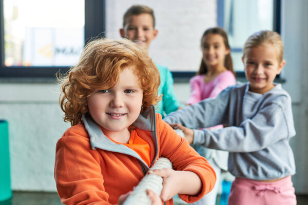 red haired cute boy pulling fitness rope alongside with his peers and smiling at camera, sport